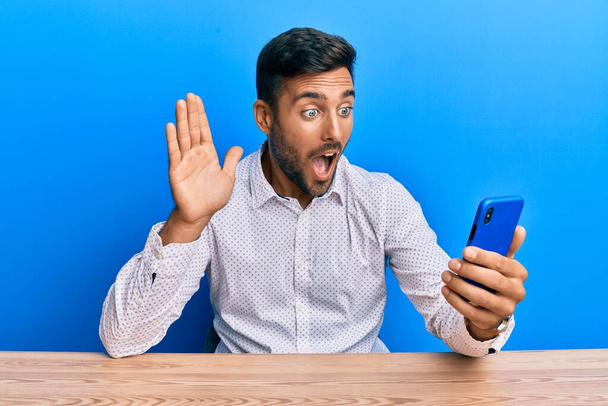 Bonito homem hispânico fazendo videochamada acenando para smartphone com medo e chocado com a expressão surpresa e surpreso, medo e rosto animado.  - Foto, Imagem