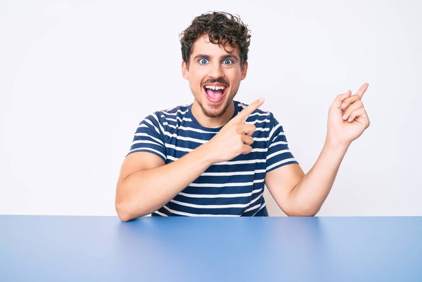 Young caucasian man with curly hair wearing casual clothes sitting on the table smiling and looking at the camera pointing with two hands and fingers to the side.  - Foto, Imagem