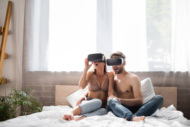excité jeune couple en utilisant vr casques tandis que assis dans le lit - Photo, image