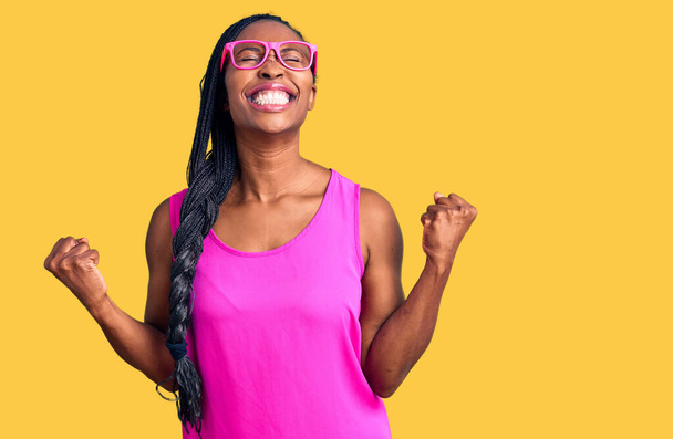 Sıradan kıyafetler ve gözlük takan genç Afro-Amerikalı kadın başarı için heyecanlanıyor kolları havada ve gözleri kapalı zaferi kutluyor. Kazanan konsept.  - Fotoğraf, Görsel