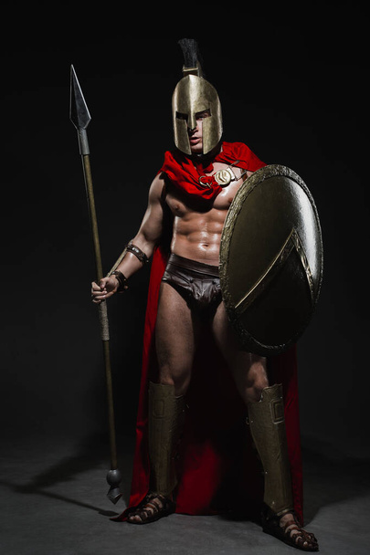 A young athletic sexy man dressed as a Roman warrior in a red cloak stands with a spear in his hand and a full-length shield on a black background. - Photo, Image