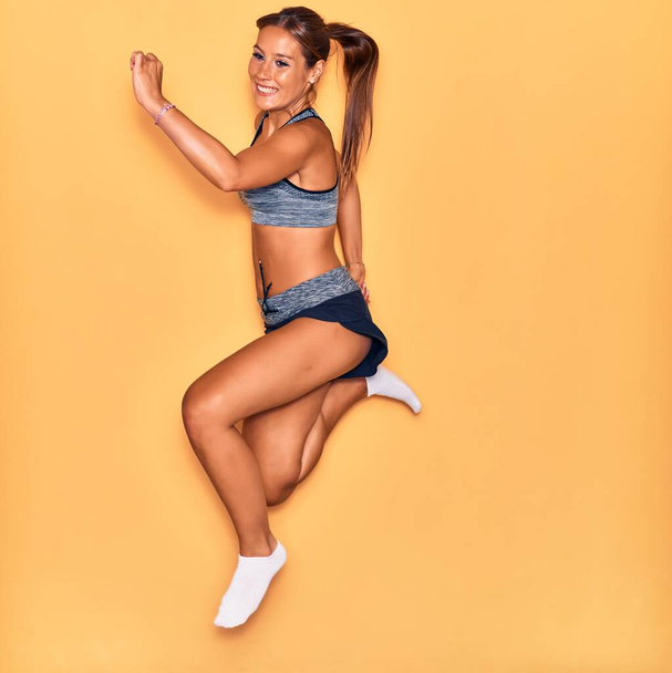 Молодая красивая латиноамериканка в спортивной одежде улыбается счастливой. Прыжки с улыбкой на лице на изолированном желтом фоне. - Фото, изображение