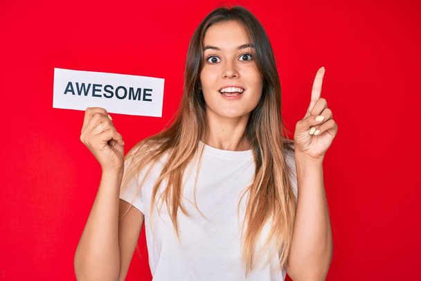 Beautiful caucasian woman holding awesome banner surprised with an idea or question pointing finger with happy face, number one  - Фото, изображение