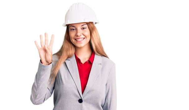Belle jeune femme portant un hardhat d'architecte montrant et pointant vers le haut avec les doigts numéro quatre tout en souriant confiant et heureux.  - Photo, image