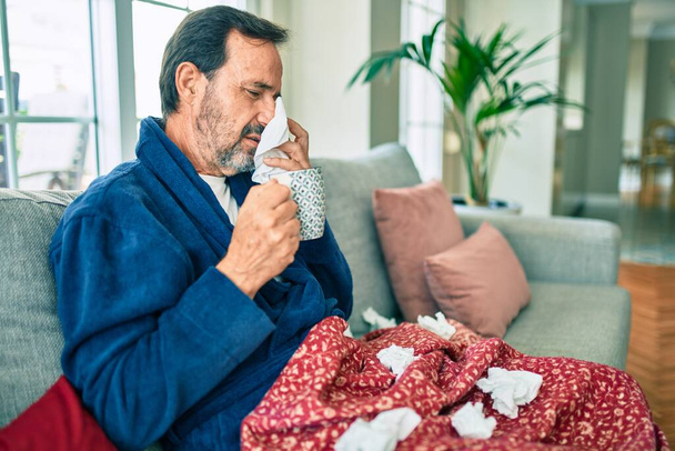 Middelbare leeftijd man voelt zich ziek met kou en koorts thuis, ziek met griepziekte zittend op de bank - Foto, afbeelding