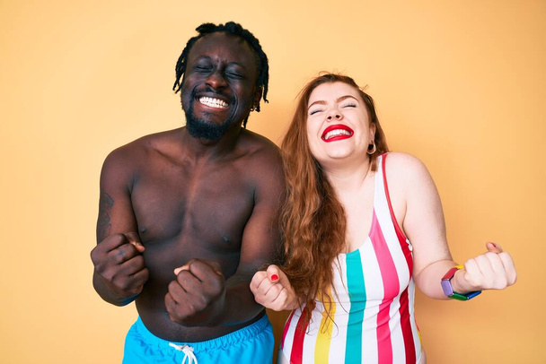水着を着た異人種間のカップルは非常に満足し、腕を上げて勝者のジェスチャーを行う興奮し、笑顔と成功のために叫んでいます。お祝いのコンセプト.  - 写真・画像