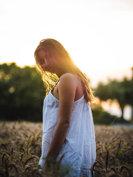  attraente femmina posa alla fotocamera nel campo di grano al tramonto. Donna caucasica con un vestito bianco in un campo di grano al tramonto. giovane caucasico felicemente sorridente nel campo - Foto, immagini