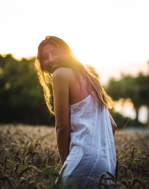  aantrekkelijke vrouw poseren aan de camera in het tarweveld bij zonsondergang. Blanke vrouw met een witte jurk in een tarweveld bij zonsondergang. jong blank gelukkig glimlachend in het veld - Foto, afbeelding