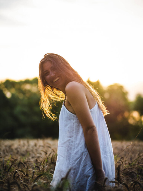 attractive female posing at the camera in the wheat field at sunset. Caucasian female with a white dress in a wheat field at sunset. young Caucasian happily smiling in the field - Фото, изображение