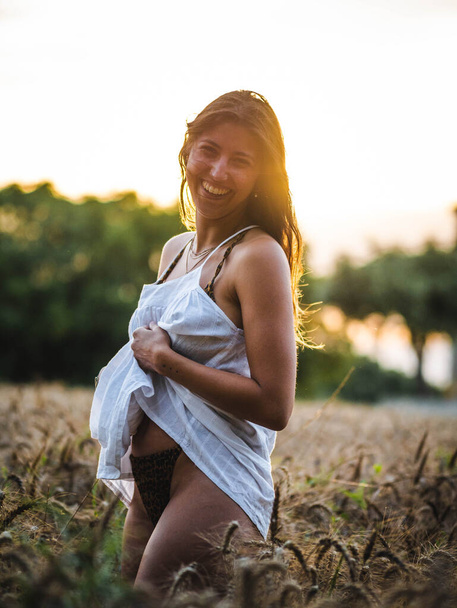  attraktive Frau posiert bei Sonnenuntergang im Weizenfeld vor der Kamera. Kaukasische Hündin mit weißem Kleid in einem Weizenfeld bei Sonnenuntergang. junge Kaukasier glücklich lächelnd auf dem Feld - Foto, Bild