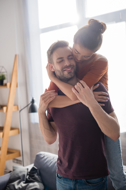 young woman hugging boyfriend from back while standing on sofa at home - Fotó, kép