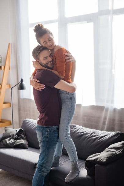 happy man hugging beloved girlfriend standing on sofa at home - Photo, image