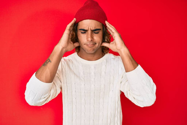 Νεαρός Ισπανός που φοράει μάλλινο πουλόβερ και χειμερινό καπέλο και υποφέρει από πονοκέφαλο απελπισμένος και αγχωμένος επειδή πονάει και ημικρανία. χέρια στο κεφάλι.  - Φωτογραφία, εικόνα