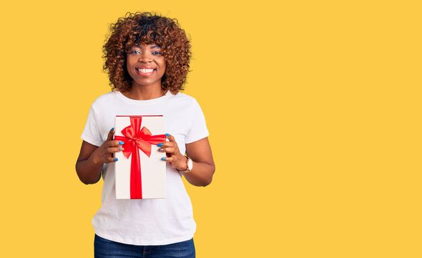 Young african american woman holding gift looking positive and happy standing and smiling with a confident smile showing teeth  - Fotó, kép