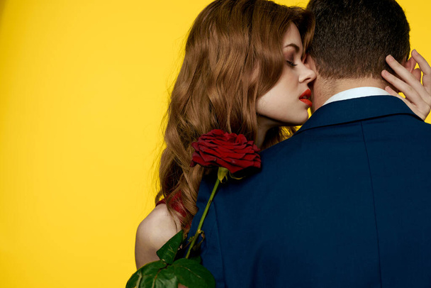 A romantic man hugs a woman in a red dress with a rose in his hand on a yellow background - Fotó, kép