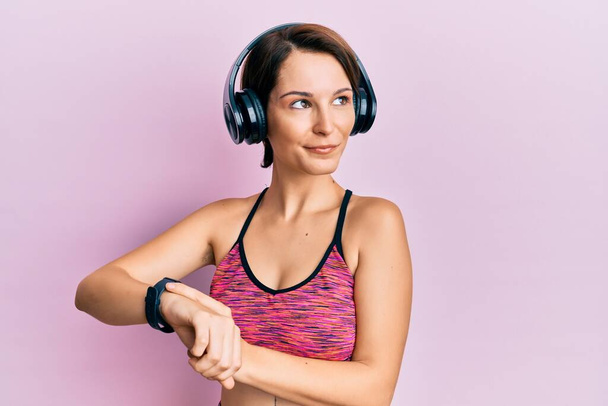 Young brunette woman with short hair using headphones and smart watch smiling looking to the side and staring away thinking.  - Foto, imagen