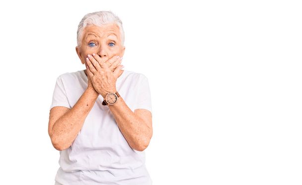 Senior mooie vrouw met blauwe ogen en grijs haar dragen casual wit tshirt geschokt bedekken mond met handen voor fout. geheim concept.  - Foto, afbeelding