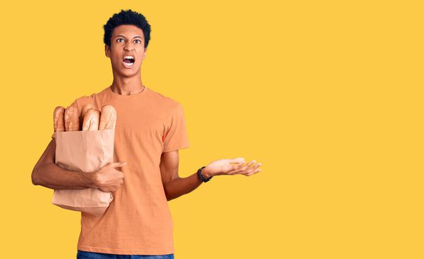 Junger afrikanisch-amerikanischer Mann hält Papiertüte mit Brot in der Hand und schreit und schreit mit aggressivem Gesichtsausdruck und erhobenen Armen. Frustrationskonzept.  - Foto, Bild