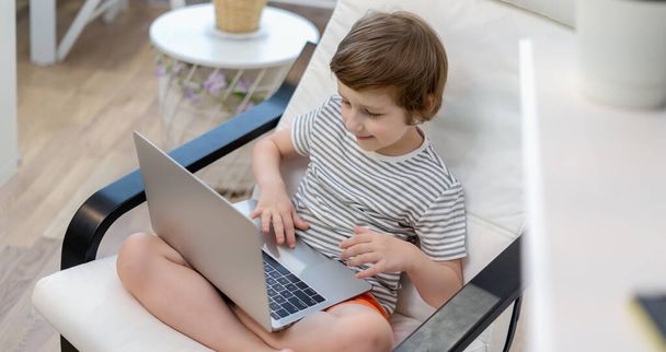 Terug naar school. Online onderwijs op afstand. Blanke glimlach kleuter jongen jongen studeren thuis met laptop notebook en huiswerk maken. Zittend op stoel met computer - Foto, afbeelding