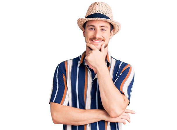 Young handsome caucasian man wearing summer hat looking confident at the camera smiling with crossed arms and hand raised on chin. thinking positive.  - Foto, immagini