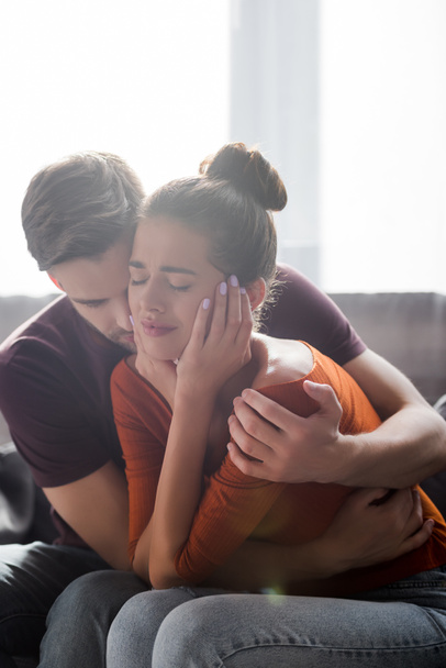 tender man hugging crying girlfriend touching face while sitting on sofa - Photo, Image