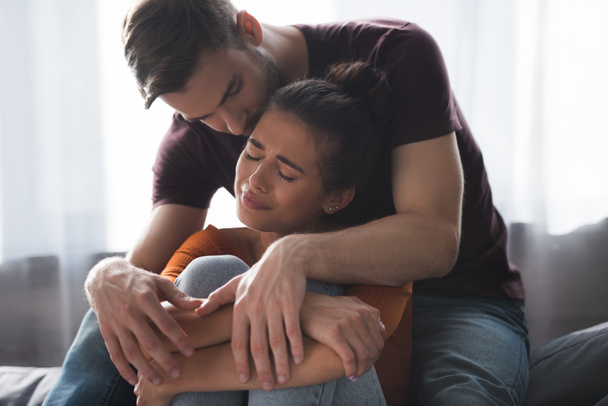tender man calming frustrated girlfriend crying on sofa at home - Photo, Image