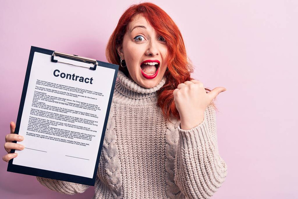 Young redhead woman holding clipboard with contract document paper over pink background pointing thumb up to the side smiling happy with open mouth - Zdjęcie, obraz
