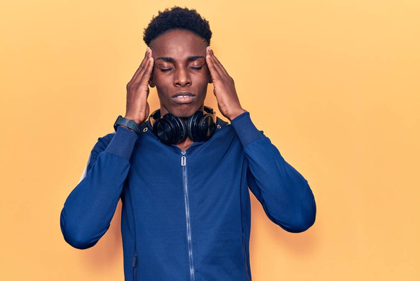 Young african american man wearing sportswear and headphones with hand on head, headache because stress. suffering migraine.  - 写真・画像