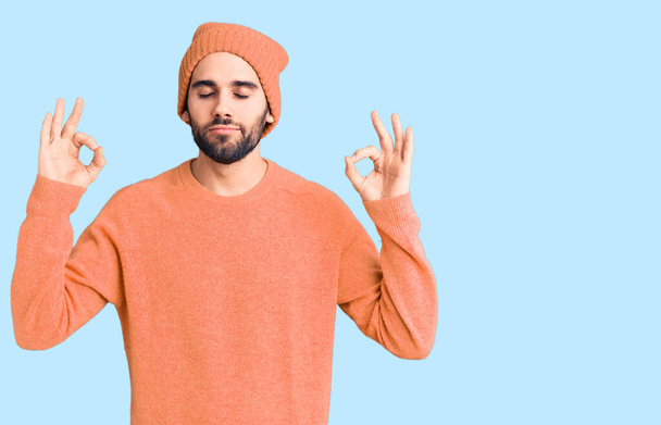 Young handsome man with beard wearing casual sweater and wool cap relax and smiling with eyes closed doing meditation gesture with fingers. yoga concept.  - Foto, imagen