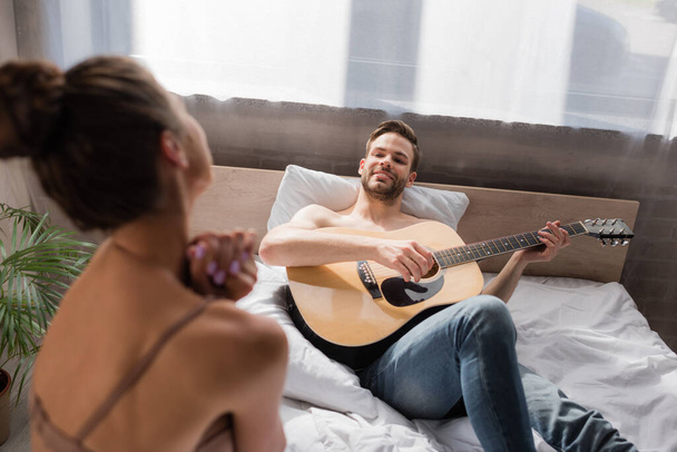 笑顔の男がベッドに横になってギターを弾きながら、彼女はぼやけた前景で踊る - 写真・画像