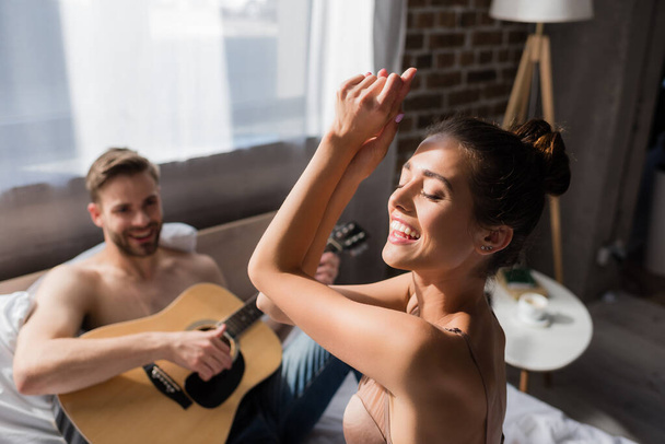 excited woman in bra dancing with closed eyes near boyfriend playing guitar on blurred background - Photo, Image