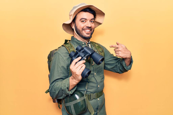 Fiatal spanyol férfi, felfedező ruhát visel, távcsövet tart, boldogan mosolyog, mutatva kezével és ujjával.  - Fotó, kép