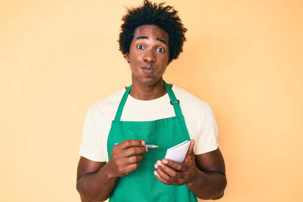 Handsome african american man with afro hair wearing waiter apron taking order puffing cheeks with funny face. mouth inflated with air, catching air.  - Photo, Image