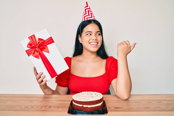 Young asian girl wearing birthday hat holding present pointing thumb up to the side smiling happy with open mouth  - Фото, изображение
