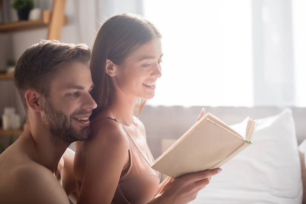 cheerful shirtless man behind seductive girlfriend reading book in bedroom - Fotó, kép