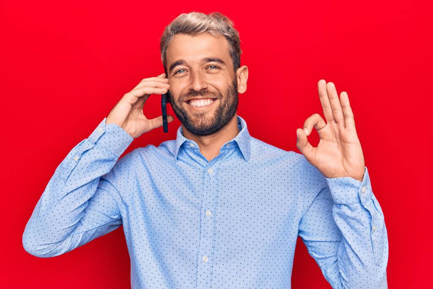 Young handsome blond man with beard having conversation talking on the smartphone doing ok sign with fingers, smiling friendly gesturing excellent symbol - Photo, Image