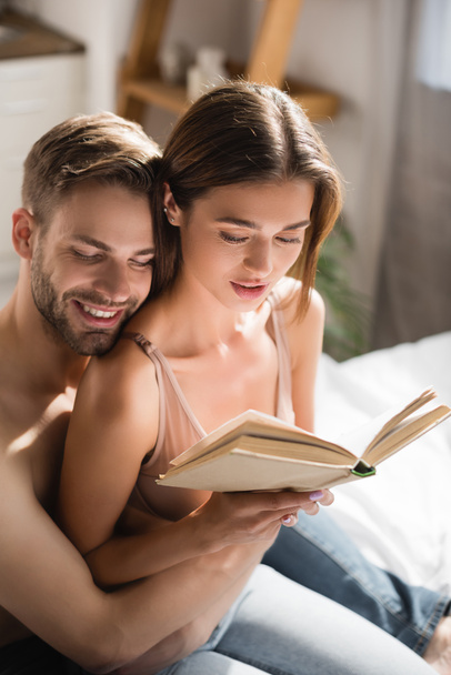happy man with closed eyes embracing sexy girlfriend reading book in bedroom - Foto, imagen