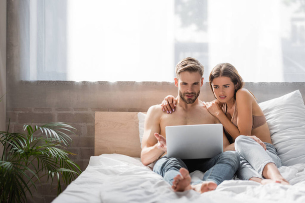 shocked woman touching shirtless boyfriend using laptop in bed - Photo, Image