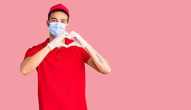 Young handsome hispanic man wearing delivery uniform and medical mask smiling in love showing heart symbol and shape with hands. romantic concept.  - Foto, Bild