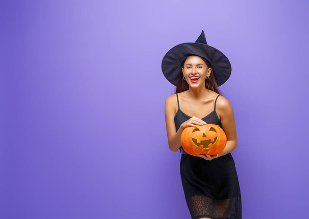 Happy Halloween! Young woman in black witch costume with pumpkin on party on purple color background. - 写真・画像
