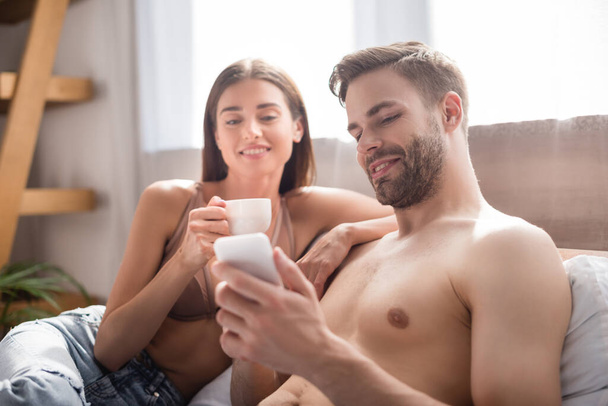 shirtless man chatting on smartphone near smiling girlfriend holding cup of coffee on blurred background - Foto, immagini