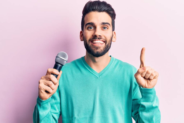 Junger gutaussehender Mann mit Bart, der ein Lied mithilfe eines Mikrofons singt und dabei mit einer Idee oder Frage lächelt, Zeigefinger mit glücklichem Gesicht, Nummer eins  - Foto, Bild
