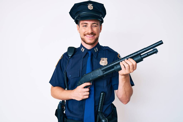 Joven hombre caucásico vistiendo uniforme policial sosteniendo escopeta guiñando el ojo mirando a la cámara con expresión sexy, cara alegre y feliz.  - Foto, imagen