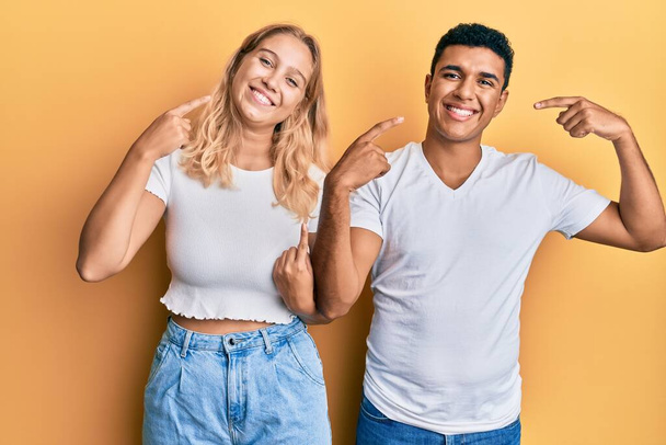 Jeune couple interracial portant un t-shirt blanc décontracté souriant et joyeux montrant et pointant du doigt les dents et la bouche. concept de santé dentaire.  - Photo, image