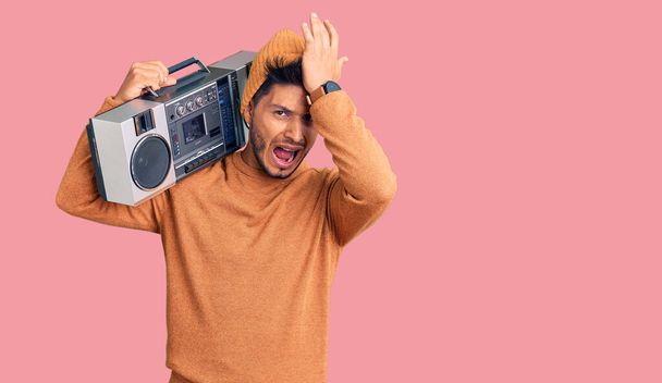 Handsome latin american young man holding boombox, listening to music surprised with hand on head for mistake, remember error. forgot, bad memory concept.  - Photo, Image