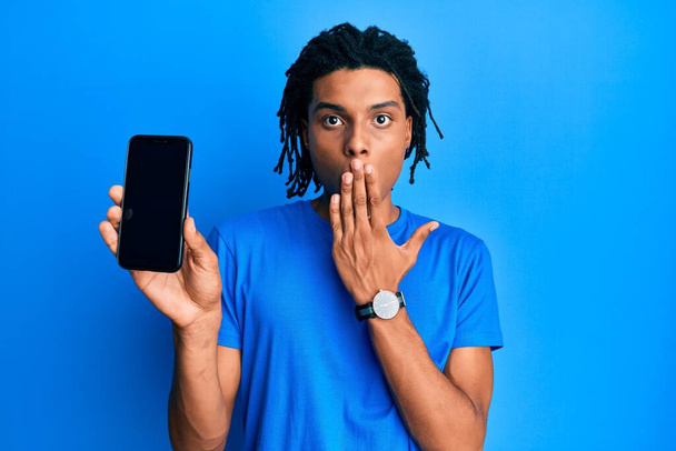 Młody, afrykański Amerykanin trzymający smartfona z pustym ekranem zakrywającym usta ręką, zszokowany i bojący się pomyłki. zaskoczona ekspresja  - Zdjęcie, obraz