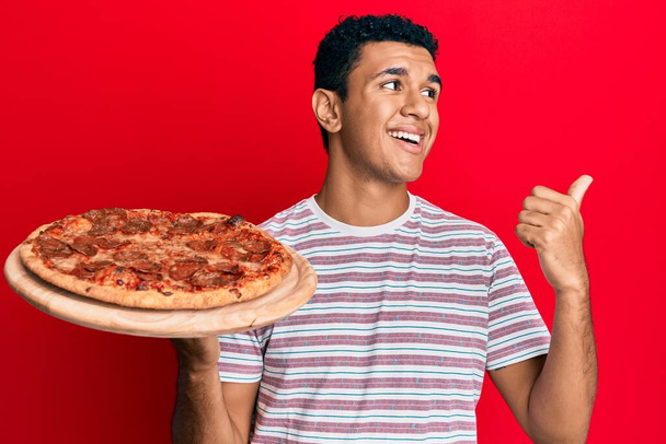 Young arab man holding italian pizza pointing thumb up to the side smiling happy with open mouth  - Photo, Image