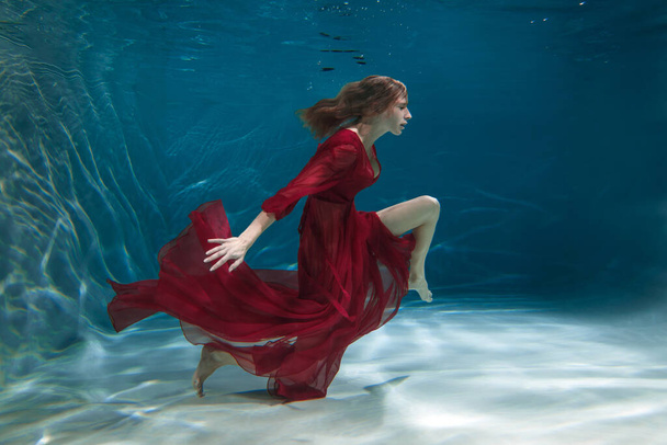 A slender girl in a long red dress swims underwater. - Zdjęcie, obraz