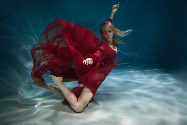 A slender girl in a long red dress swims underwater. - 写真・画像