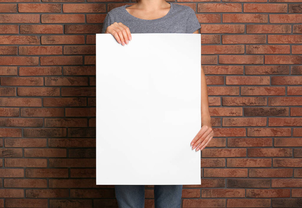 Woman holding white blank poster near red brick wall, closeup. Mockup for design - Φωτογραφία, εικόνα